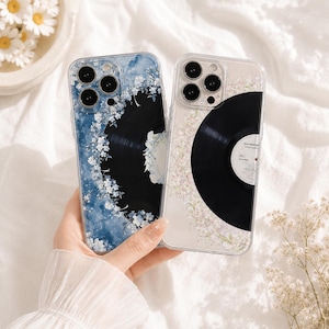 Puede incluir: Dos fundas transparentes para smartphone con un gráfico de disco de vinilo negro. La funda izquierda tiene un fondo de acuarela azul con detalles florales blancos. La derecha es transparente con una corona floral blanca alrededor del disco.