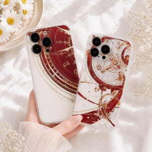 Può includere: Due cover trasparenti per smartphone con un design celeste in rosso intenso e oro. La cover sinistra mostra una carta stellare circolare parziale, mentre quella destra presenta intricati ingranaggi meccanici e linee dorate su sfondo bianco.