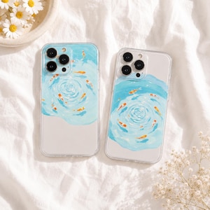 May include: Two clear phone cases featuring a painted design of orange koi fish swimming in light blue water ripples. The cases have cutouts for the camera lenses and a transparent lower section, showcasing a serene aquatic pattern.
