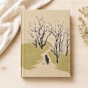 May include: A hardcover book with a tan fabric cover featuring a printed illustration of a black cat walking along a path through a green meadow with two bare trees adorned with small white blossoms against a light beige background.