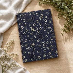 May include: A rectangular hardcover journal featuring a dark navy blue cover with a repeating pattern of small cream-colored cherry blossoms and stylized bamboo stalks. The book is oriented vertically and has a clean, flat, and uniform surface.