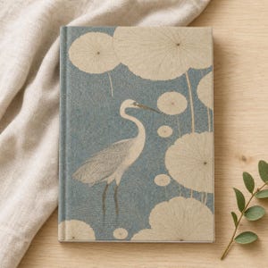May include: A hardcover journal featuring a blue background with a textured circular pattern. The cover displays a white heron standing among several large, pale beige circular lily pad shapes. The design is printed on the front of the notebook.