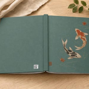 May include: A teal hardcover book featuring an illustration of two koi fish swimming in water with scattered red maple leaves. The back cover displays a small white square QR code near the spine, and the spine has a small brown rectangular mark.