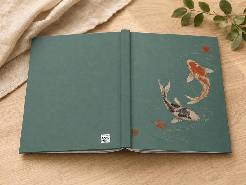 May include: A teal hardcover book featuring an illustration of two koi fish swimming in water with scattered red maple leaves. The back cover displays a small white square QR code near the spine, and the spine has a small brown rectangular mark.