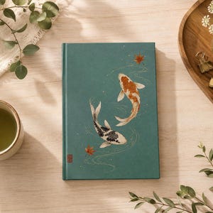 May include: A rectangular hardcover journal with a teal cover featuring an illustration of two koi fish swimming in water with gold line accents and small red maple leaves. A small red square seal is printed in the bottom left corner of the cover.