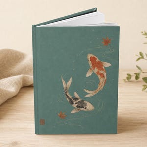 May include: A hardcover book with a matte teal cover featuring an illustration of two koi fish swimming in water with scattered red maple leaves. One koi is orange and white, while the other is black and white. The book is slightly open at the top.