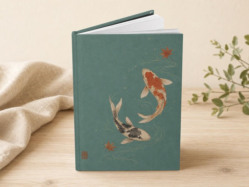 May include: A hardcover book with a matte teal cover featuring an illustration of two koi fish swimming in water with scattered red maple leaves. One koi is orange and white, while the other is black and white. The book is slightly open at the top.