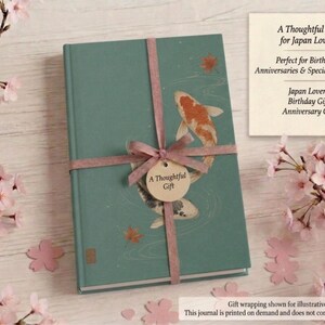 May include: A teal hardcover journal featuring an illustration of two koi fish swimming in water with scattered maple leaves. The journal is tied with a mauve ribbon and a circular tag that reads A Thoughtful Gift in the center of the cover.