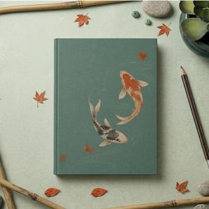 May include: A rectangular hardcover journal with a muted sage green cover featuring an illustration of two koi fish swimming in a circle, one orange and white and the other black and white, surrounded by small scattered orange maple leaves.