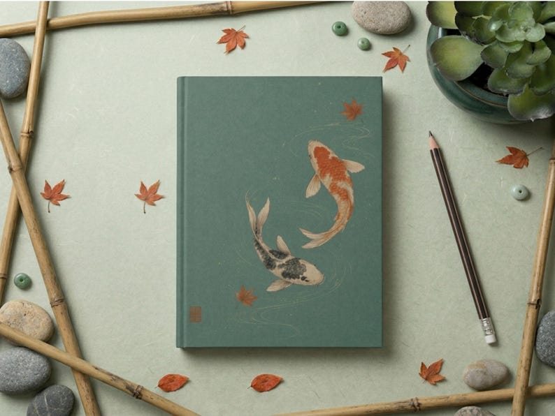 May include: A rectangular hardcover journal with a muted sage green cover featuring an illustration of two koi fish swimming in a circle, one orange and white and the other black and white, surrounded by small scattered orange maple leaves.
