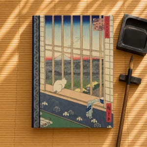 May include: A rectangular notebook featuring a Japanese woodblock print of a white cat sitting on a windowsill overlooking a landscape with Mount Fuji in the distance. The cover includes vertical window panes, a blue patterned border, and red text.