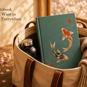 May include: A teal notebook featuring two koi fish and maple leaves, placed inside a beige canvas tote bag with brown leather handles. The notebook cover displays the text A Notebook You'll Want to Carry Everywhere. A black water bottle is also inside.