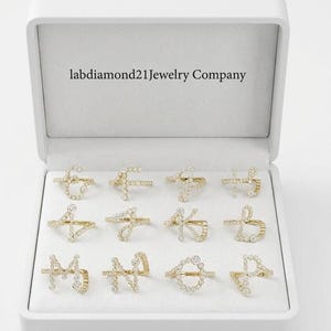 Op de afbeelding: Een witte sieradendoos met twaalf goudkleurige initialenringen, elk bezet met kleine, sprankelende diamanten. De ringen zijn netjes in de doos gerangschikt, met de bedrijfsnaam "labdiamond21Jewelry Company" op het binnendeksel.