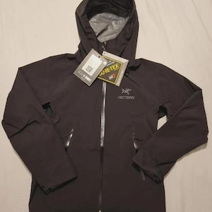 Può includere: Una giacca nera con cappuccio Arc'teryx dotata di cerniera frontale, maniche lunghe e due tasche laterali con zip. La giacca presenta un'etichetta del marchio Gore-Tex attaccata sul petto e un logo bianco ricamato sulla parte superiore sinistra.