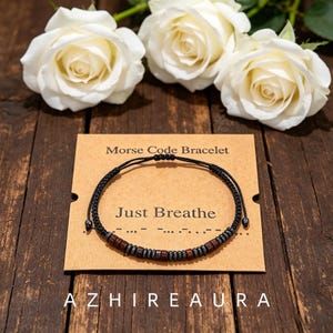 Könnte beinhalten: Ein schwarzes Kordelarmband mit abwechselnden dunkelbraunen Holzperlen und kleinen metallisch grauen Perlen in einem Morsecode-Muster. Das Armband liegt auf einer beigen Karte mit der Aufschrift Morse Code Bracelet und Just Breathe.