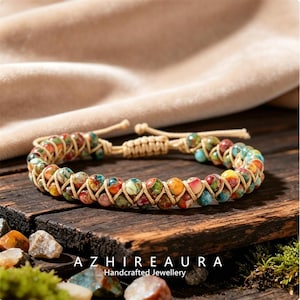 Puede incluir: Un brazalete artesanal con cuentas coloridas y estampadas. Las cuentas están ensartadas con un cordón beige, creando un efecto tejido. El brazalete se exhibe sobre una superficie de madera. El nombre de la marca "AZHIRE AURA" es visible.