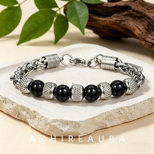 May include: A bracelet featuring a silver-toned wheat chain, alternating with smooth black spherical beads and textured silver-toned cylindrical spacers. The piece includes a lobster claw clasp and a small metal tag. The text AZHIEAURA is visible.
