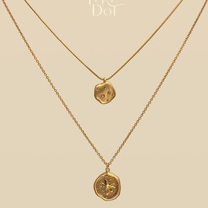 May include: Two gold necklaces with different pendants. The top necklace has a textured, irregular pendant with small, clear stones. The bottom necklace features a circular pendant with a bee design. The necklaces are displayed against a neutral background, with the brand name "Peri Dot" in the upper right.