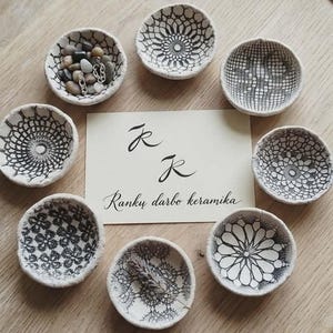 Puede incluir: Una colección de pequeños cuencos de cerámica hechos a mano con intrincados diseños negros similares al encaje. Los cuencos están dispuestos sobre una superficie de madera, algunos llenos de piedras y joyas. Una tarjeta con caligrafía dice "Ranku darbo keramika".