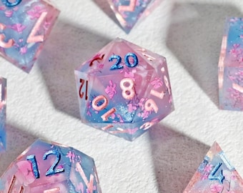 Op de afbeelding: Een set van zeven polyhedrale dobbelstenen voor tabletopspellen. Elke dobbelsteen is een doorschijnende hars met een roze en blauw bloemenontwerp en blauwe nummering. De dobbelstenen bevatten een d4, d6, d8, d10, d12, d20 en een percentieldobbelsteen.