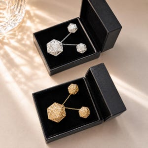 May include: Two black jewellery boxes containing polyhedral dice sets. One box holds a silver-toned set with a large twenty-sided die and two smaller dice connected by chains. The other box contains a gold-toned set with identical dice configurations.