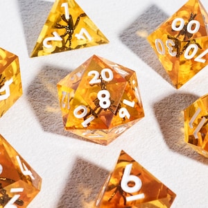 May include: A set of seven polyhedral dice made of translucent amber-colored resin, each containing a small black insect figure embedded inside. The dice feature crisp white numbers engraved on their surfaces, including a d20, d12, d10, d8, d6, and d4.