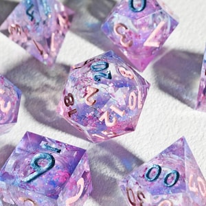 May include: A set of seven polyhedral dice in shades of purple, pink, and blue. The dice are transparent with glitter and swirls inside, and the numbers are in a contrasting color. The dice are for tabletop games.