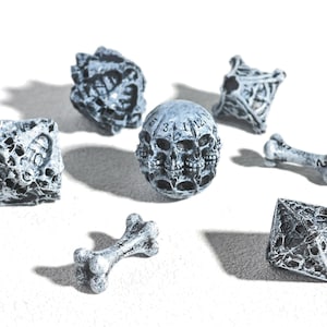 Könnte beinhalten: Ein Set aus sieben polyedrischen Spielwürfeln und knochenförmigen Markern mit einer verwitterten, steingrauen Oberfläche. Die Sammlung umfasst verschiedene geometrische Formen mit komplexen Totenkopf- und Gitterstrukturen sowie schwarzen Zahlen.
