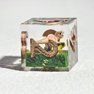 May include: A clear resin die with a miniature dinosaur scene inside. The die features copper-colored numbers and a small dinosaur figurine on a green base. The die is cube-shaped and has a detailed, artistic design.