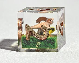 Puede incluir: Un dado de resina transparente con una escena de dinosaurios en miniatura en su interior. El dado presenta números de color cobre y una pequeña figura de dinosaurio sobre una base verde. El dado tiene forma de cubo y tiene un diseño artístico detallado.