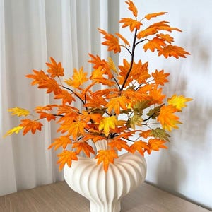 Puede incluir: Un jarrón de cerámica blanca con forma redondeada y acanalada contiene una rama de hojas de arce artificiales de color naranja y amarillo hechas con limpiapipas. El texto "Video Tutorial: Pipe Cleaner Maple Leaves" aparece en la esquina inferior derecha.