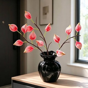 Op de afbeelding: Een glanzend zwarte keramische vaas met een geschulpte rand bevat verschillende donkerbruine takken met kleine, druppelvormige roze en witte bloemen. De vaas heeft een afgerond, geribbeld lichaam en staat op een vlak oppervlak.