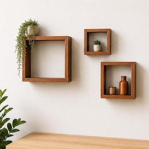 May include: Three square wooden wall shelves in a medium brown finish. The largest shelf holds a trailing green plant in a pot. The top smaller shelf contains a small succulent in a pot, and the bottom shelf holds a brown vase and a small cup.