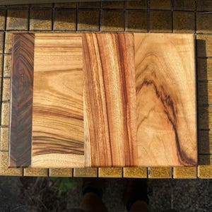 Op de afbeelding: Een rechthoekige houten snijplank opgebouwd uit vier afzonderlijke verticale houten panelen. Het paneel aan de uiterste linkerzijde is donkerbruin met een fijne nerf, terwijl de overige drie panelen lichtere bruine en goudbruine tinten hebben met opvallende, wervelende houtnerfpatronen.