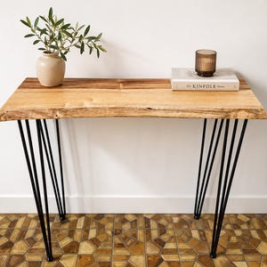 May include: A rectangular console table with a natural live-edge light wood top and four black metal hairpin legs. On the surface sits a beige ceramic vase with green leaves and a book titled The Kinfolk Home with a brown glass candle on top.