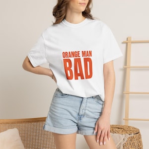 Könnte beinhalten: Weißes T-Shirt mit den Worten "ORANGE MAN BAD" in kräftigen orangefarbenen Buchstaben. Das Shirt wird mit Jeansshorts getragen. Das Model steht vor einem hellen Hintergrund.
