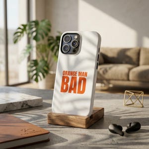 Könnte beinhalten: Eine weiße Smartphone-Hülle mit dem Text ORANGE MAN BAD in fetten orangefarbenen Buchstaben. Die Hülle hat eine Aussparung für ein Triple-Kamera-System und steht aufrecht auf einem kleinen Holzständer auf einer flachen Oberfläche.