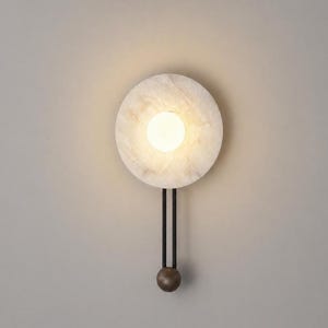 May include: A wall sconce featuring a circular, translucent white stone backplate with a central glowing orb light. Two thin, vertical black metal rods extend downward from the backplate, connecting to a small, dark, spherical metal weight at the base.