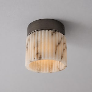 Op de afbeelding: Een plafondlamp met een cilindrische, geribbelde kap van wit marmer met grijze aders. De bovenkant van het armatuur is afgewerkt met een donkere, geborstelde, ronde metalen houder die de stenen kap op zijn plaats houdt.