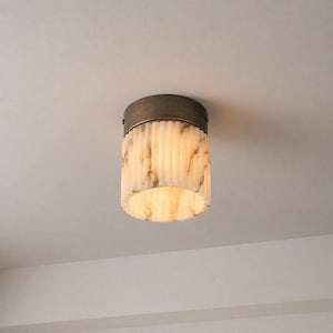 May include: A cylindrical ceiling light fitting with a bronze-coloured metal base and a textured, off-white shade. The shade has vertical lines and a marbled appearance. The light is on, illuminating the room.