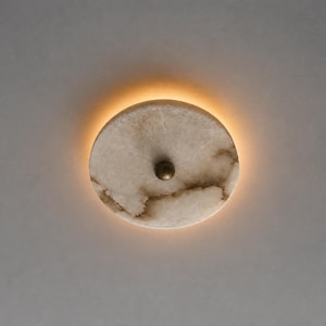 May include: A round, flush-mount ceiling light fixture. The light has a circular, off-white marble shade with brown veining and a small, brass-colored knob in the center. A warm, orange glow emanates from behind the shade.