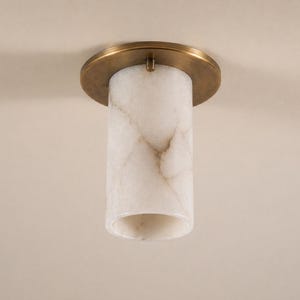 Op de afbeelding: Een cilindrische plafondlamp met een messingkleurige cirkelvormige basis. De lampenkap is gemaakt van een wit, marmerachtig materiaal met bruine aderen. De armatuur is gemonteerd op een neutraal gekleurd plafond.