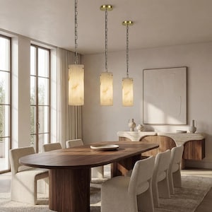 May include: Three pendant lights with cylindrical shades hang above a wooden dining table. The shades are a cream color with a vertical ribbed texture and a warm glow. The dining room features a large oval table, upholstered chairs, and a wooden sideboard.