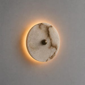 May include: A round, off-white wall sconce with a warm, glowing light. The sconce features a marble-like pattern with brown veining and a small, brass-colored knob in the center. The light source is a soft, diffused glow around the edge of the sconce.