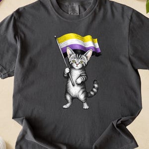 May include: Dark gray t-shirt featuring a detailed illustration of a gray tabby cat holding a non-binary pride flag. The flag is yellow, white, purple, and black. The shirt is displayed on a light brown surface with white sneakers and a rainbow flag.