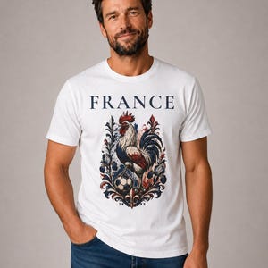 May include: White t-shirt with the word "FRANCE" in navy blue. The shirt features a detailed graphic of a rooster, a soccer ball, and floral elements in red, white, and blue. The design is centered on the front of the shirt.
