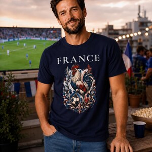 May include: Navy blue t-shirt with the word "FRANCE" above a detailed graphic of a rooster, soccer ball, and floral elements in red, white, and blue. The shirt is worn by a person in blue jeans.