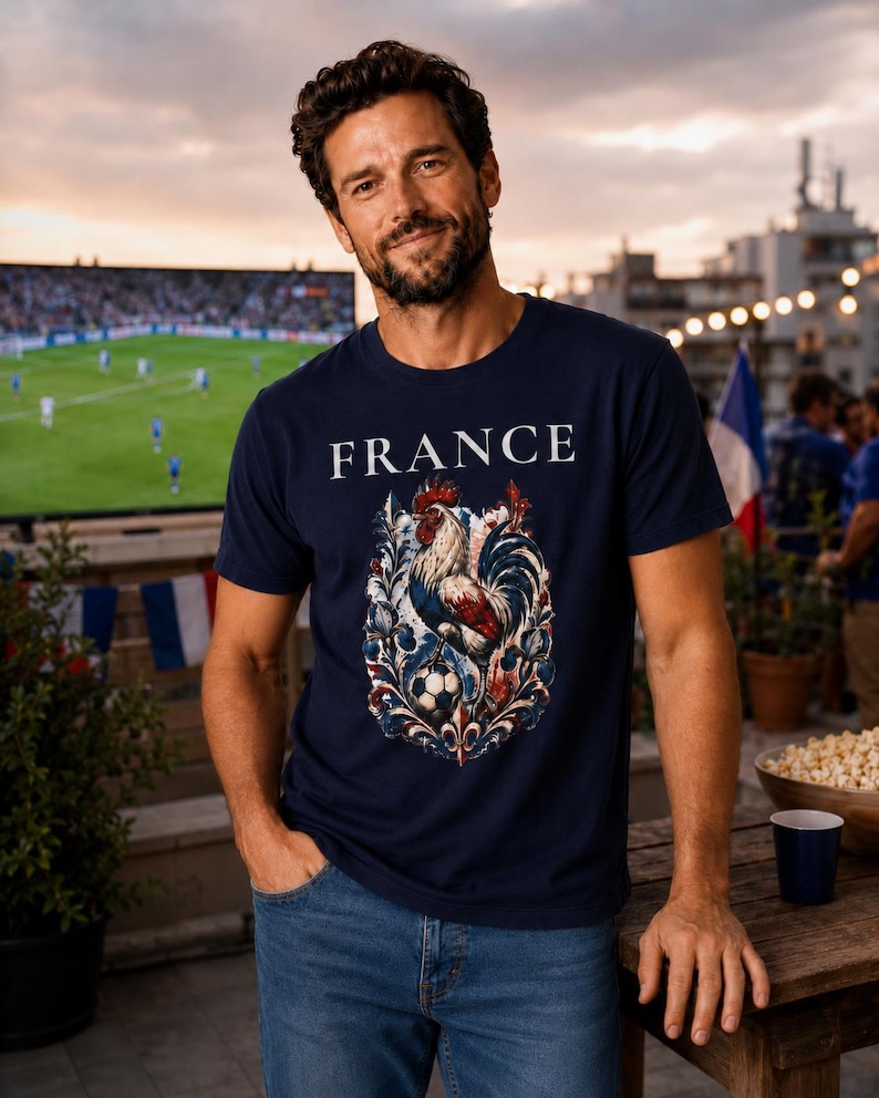 May include: Navy blue t-shirt with the word "FRANCE" above a detailed graphic of a rooster, soccer ball, and floral elements in red, white, and blue. The shirt is worn by a person in blue jeans.