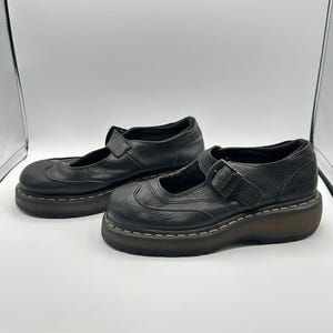 May include: A pair of black leather Mary Jane shoes featuring a textured finish, a rounded toe with decorative stitching, and an adjustable strap with a buckle. The shoes are set on a thick, dark brown platform sole with visible yellow stitching.
