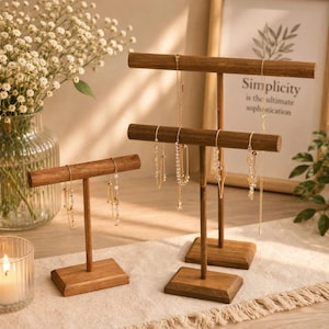 May include: Two wooden jewelry display stands with rectangular bases and horizontal bars. The taller stand has two tiers, while the shorter one has one tier. Several gold-toned necklaces and bracelets with small beads and charms hang from the bars.